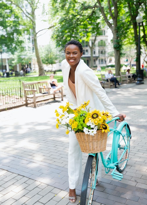 Smiling, dark-skinned woman wearing an off-white suit and holding a turquoise bike with a basket of sunflowers