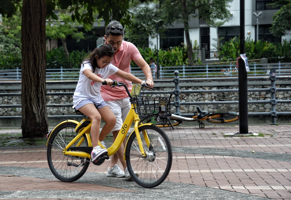 Man in a peach shirt helping a young girl on a yellow bike learn to ride