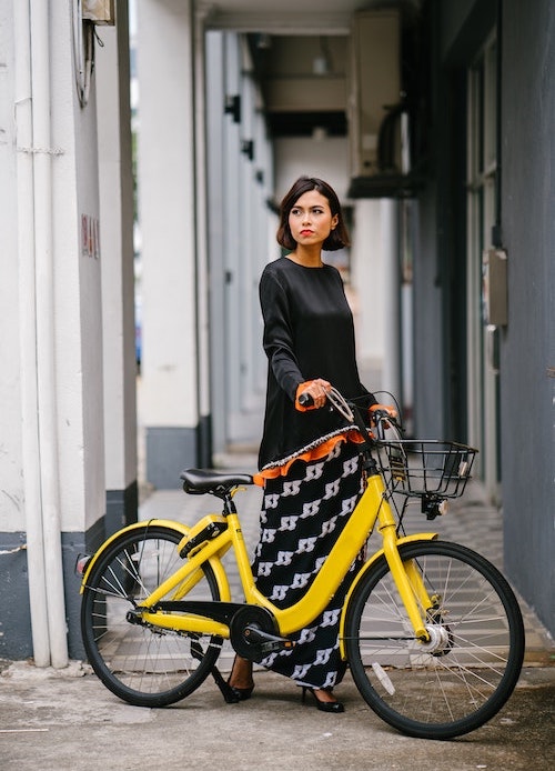 Chinese woman looking to her right, wearing a black tunic and black and white graphic print skirt standing behind a yellow e-bike
